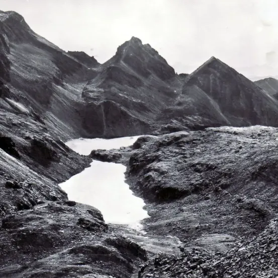 Ein Schwarz-Weiß-Bild einer Bergregion mit einem schmalen, fließenden Fluss und felsigem Gelände. Der Himmel ist bewölkt.