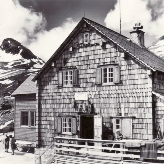 Ein Schwarz-Weiß-Foto eines Holzhauses mit steilem Dach und Schornstein, gelegen in einem Berggebiet. Eine Frau sitzt auf einer Veranda, und ein Mann und eine Frau stehen in der Nähe des Hauses.