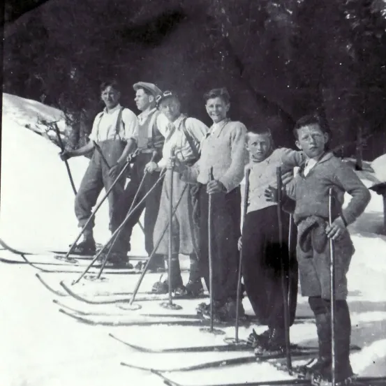 Ein Schwarz-Weiß-Foto zeigt eine Gruppe junger Männer, die im Schnee mit Skiern stehen und möglicherweise für ein Foto posieren. Sie tragen alle Skianzüge, Mützen und halten Skistöcke.