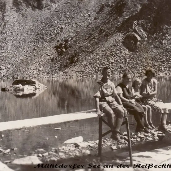 Schwarz-Weiß-Foto von vier Menschen, die auf einer Bank am See sitzen, mit einem Berg im Hintergrund.