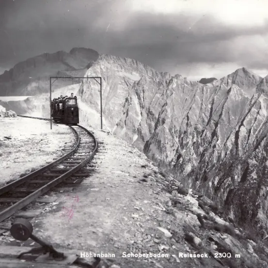 Ein Schwarz-Weiß-Bild zeigt einen Bergbahnzug, der eine steile Bergstrecke hinauffährt, mit einem bewölkten Himmel darüber.