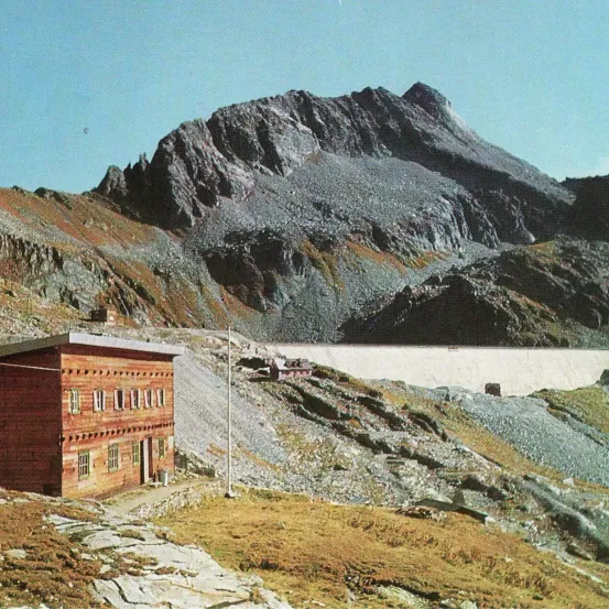 Ein kleines Holzgebäude steht am Hang eines Berges, in der Nähe eines Damms. Felsige Hänge umgeben das Gebäude, und ein großer Berg ist im Hintergrund.