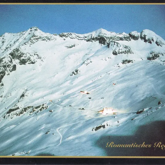 Luftaufnahme einer verschneiten Bergkette mit klarem blauem Himmel darüber. Eine Skipiste ist sichtbar.