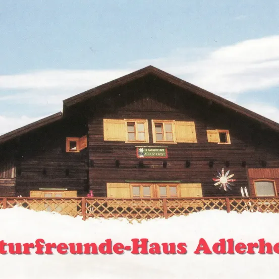 Ein Chalet-Stilgebäude mit gelben Fensterläden und einem Schild, das 'TV Naturfreunde Adlerhorst' liest, bedeckt mit Schnee und einem Zaun davor.