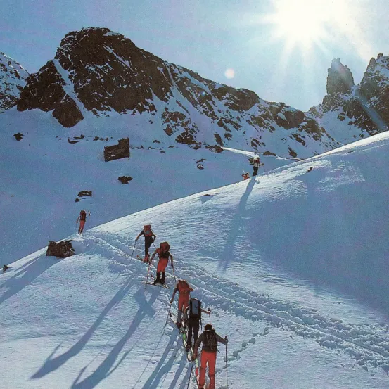 Eine Gruppe von Skifahrern in roter Ausrüstung klettert einen verschneiten Berg unter einem strahlenden Sonnenlicht. Schatten werden auf den Schnee geworfen.