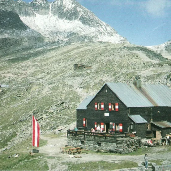 Eine Berghütte mit einer Flagge und Leuten, die auf der Veranda sitzen, umgeben von felsigem Gelände und schneebedeckten Bergen im Hintergrund.