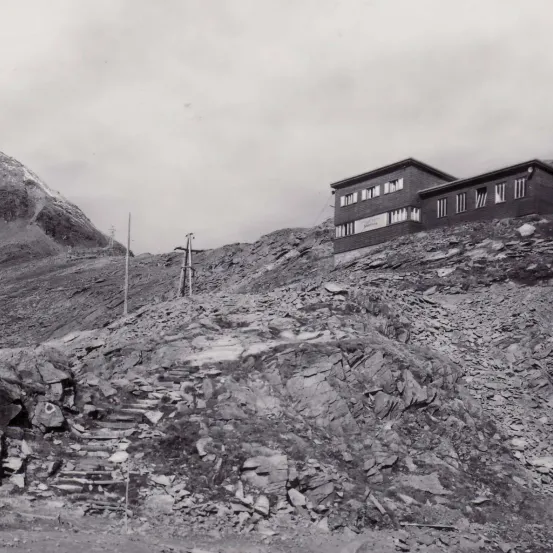 Ein Schwarz-Weiß-Foto eines felsigen Berghangs mit einem Gebäude rechts. Treppen führen zum Gebäude hinauf.