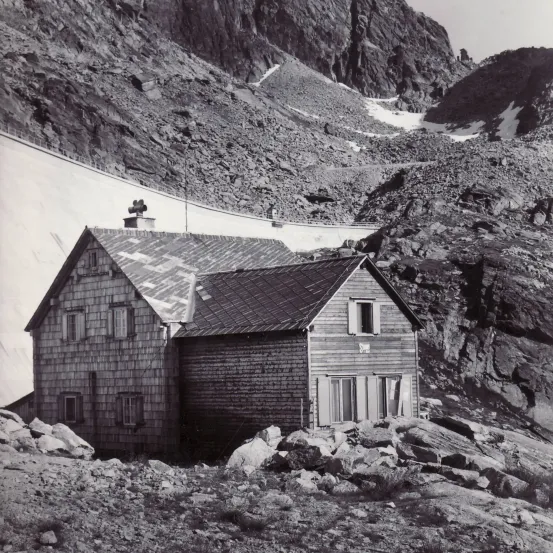 Eine Schwarz-Weiß-Aufnahme eines Holzhauses auf einem felsigen Berg, mit schneebedeckten Bergen im Hintergrund.