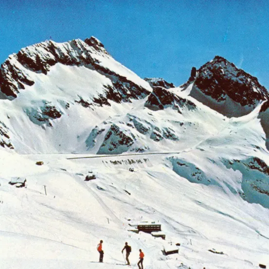 Drei Skifahrer sind auf einem verschneiten Berg mit felsigen Gipfeln. Sie fahren den Hang hinunter, einer trägt einen roten Mantel.