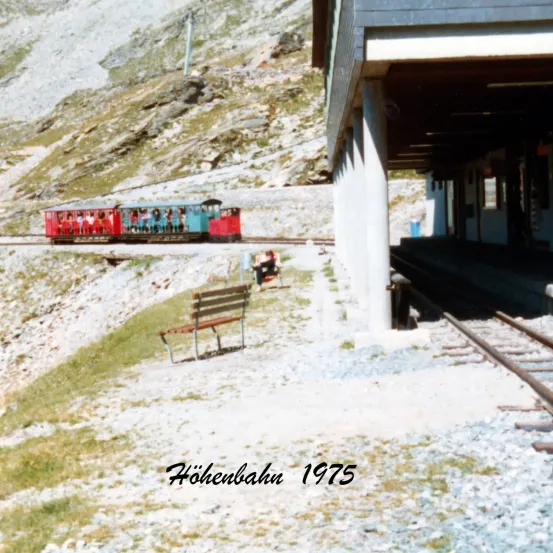 Das Bild zeigt einen roten und blauen Zug auf Gleisen, mit Menschen im Inneren. Die Gleise sind von Felsen und Gras umgeben. Eine Bank befindet sich neben den Gleisen. Der Bahnhof hat ein Dach und Säulen. Das Bild ist mit 'Hohenbahn 1975' beschriftet.