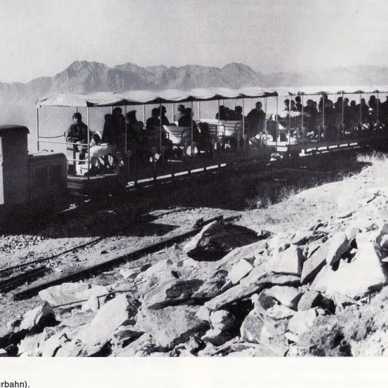 Ein Schwarz-Weiß-Foto eines Zuges mit Menschen, die darauf fahren, auf einer Bergstrecke mit Felsen an der Seite.