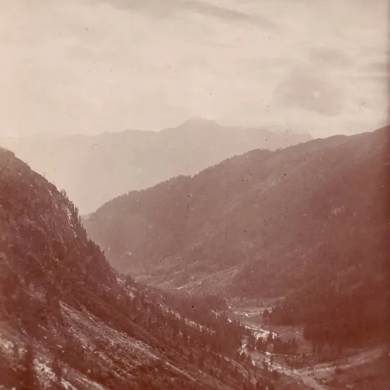 Ein sepiafarbenes Foto zeigt ein Tal mit steilen, bewaldeten Bergen. Ein schmaler Fluss schlängelt sich durch die Talsenke. Der Himmel ist bedeckt.