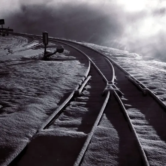 Ein Schwarz-Weiß-Bild zeigt eine verschneite Landschaft mit zwei parallelen Bahngleisen, möglicherweise auf einem Berg, bedeckt mit Schnee.