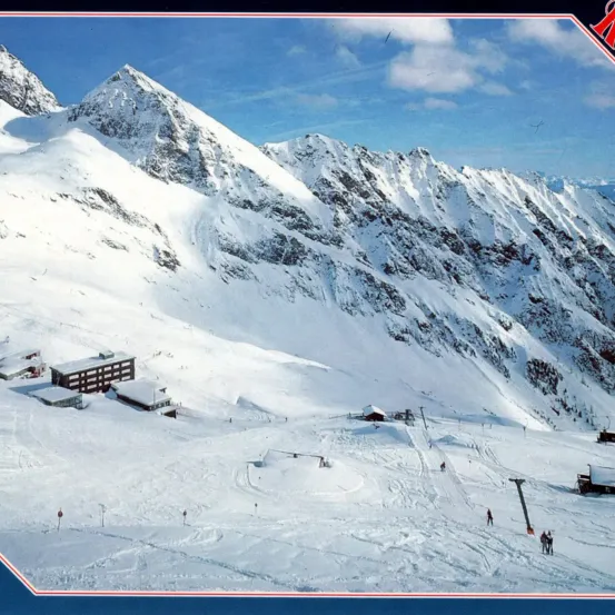 Eine verschneite Berglandschaft mit Skifahrern und Skiliften. Gebäude sind im Schnee eingebettet. Der Himmel ist blau mit einigen Wolken.