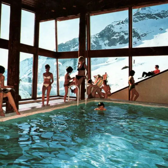 Eine Gruppe von Menschen in Badekleidung ist um einen Pool herum, mit einem verschneiten Berg im Hintergrund. Einige schwimmen, während andere sitzen oder am Poolrand stehen.
