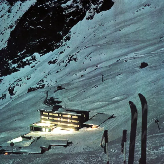 Eine Skihütte mit beleuchteten Fenstern steht auf einem verschneiten Berghang. Im Vordergrund sind Skier und Stöcke zu sehen.