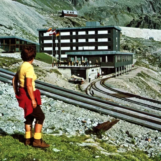 Eine Frau steht am Hang eines Berges mit einer Eisenbahnstrecke vor ihr und blickt auf ein großes Gebäude mit mehreren Fahnen auf Stangen.