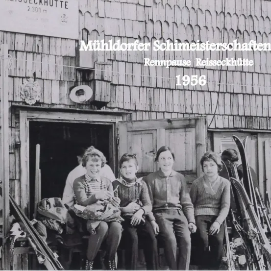 Eine Gruppe von Menschen sitzt 1956 vor einem Holzgebäude mit Skiern. Das Gebäude hat ein Schild mit der Aufschrift Muhldorfer Schimeisterschaft.