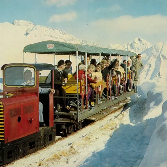 Ein Oldtimer-Zug mit grünem Dach und überdachten Sitzen transportiert Passagiere durch verschneite Berge. Einige Leute stehen und halten sich an den Geländern fest. Der Fahrer sitzt in der Kabine.