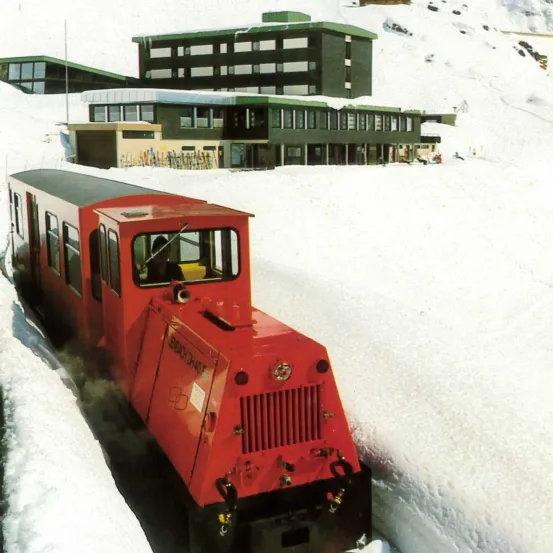 Ein roter Zug bewegt sich durch eine verschneite Landschaft mit einem großen grünen Gebäude im Hintergrund.