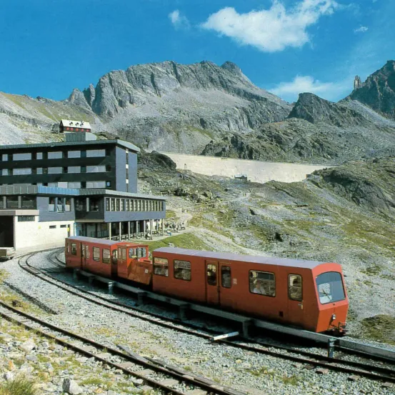 Ein roter Zug fährt auf einer Schotterstrecke in einer Gebirgsregion, mit einem Gebäude und einem Damm im Hintergrund.