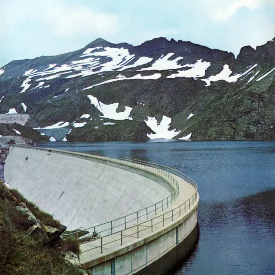 Ein Betondamm ist quer über einen See gebaut, mit schneebedeckten Bergen im Hintergrund. Der Damm hat eine gekrümmte Oberfläche und ein Metallgeländer. Der See ist ruhig mit einer Reflexion des Himmels.