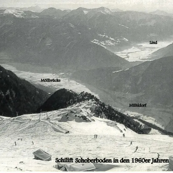 Eine Schwarz-Weiß-Fotografie einer Berglandschaft aus den 1960er Jahren. Es zeigt eine Skiliftanlage namens 'Schilift Schoberboden' mit Bergen namens 'Lind', 'Möllbrücke' und 'Mühldorf'. Es sind Skifahrer auf den Hängen zu sehen.