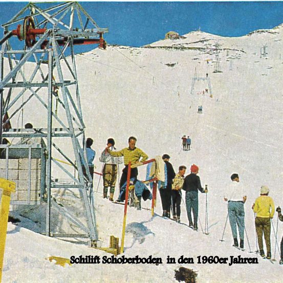 Ein Vintage-Foto zeigt Menschen am Skilift Schoberboden in den 1960er Jahren, gekleidet in Retro-Skiausrüstung, mit einem verschneiten Berg im Hintergrund.