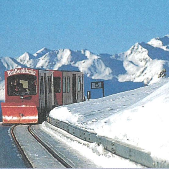 Ein roter Zug auf verschneiten Gleisen mit 'Reil3eckbahn' drauf, der sich in Richtung verschneiter Berge mit einem Schild '5' bewegt.