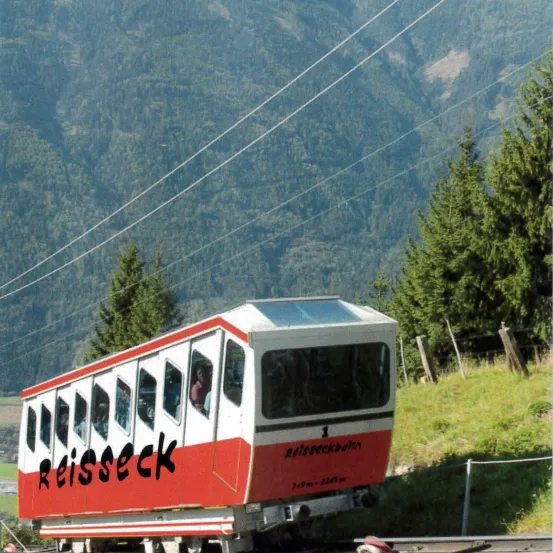 Ein rot-weißer Zug mit der Aufschrift 'Reisseck' fährt einen Berg hinauf, mit Bäumen und Stromleitungen im Hintergrund.