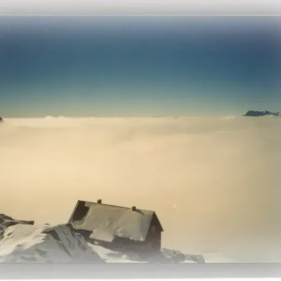 Eine Hütte in einem verschneiten Gebirgsgebiet, bedeckt von Nebel, mit schneebedeckten Gipfeln in der Ferne unter einem klaren Himmel.