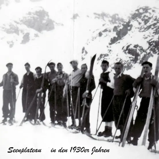 Eine Gruppe junger Männer in Winterkleidung, die in einem verschneiten Bereich mit Bergen im Hintergrund steht. Sie halten Skier und scheinen für ein Foto zu posieren. Das Foto stammt aus den 1930er Jahren.