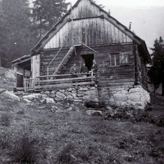Ein Schwarz-Weiß-Foto zeigt drei Männer auf der Veranda einer Holzblockhütte. Die Hütte ist von Steinen und Gras umgeben.
