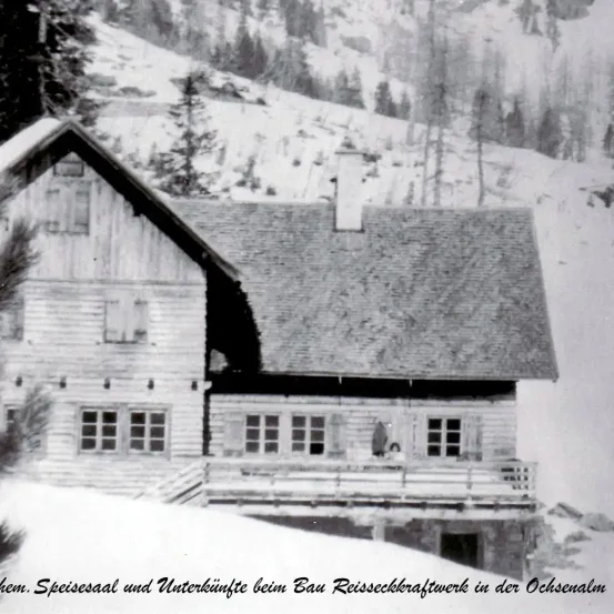 Ein Schwarz-Weiß-Foto zeigt ein rustikales Holzhaus in einem verschneiten Gebirgsgebiet, mit einer Person auf der Veranda stehend.