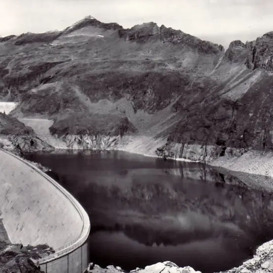 Ein Schwarz-Weiß-Foto eines Staudamms in einem Gebirgsgebiet mit einem Gewässer, das die umliegende Landschaft reflektiert.