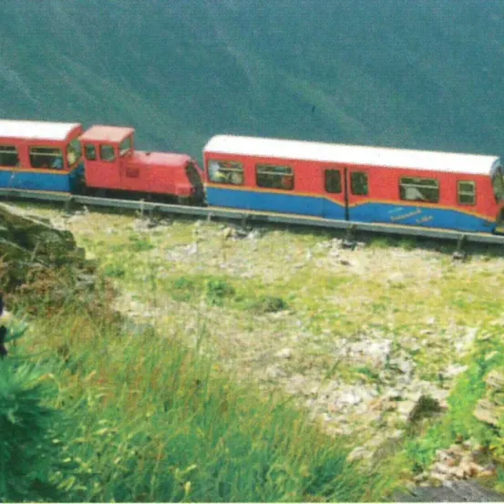 Ein rot-blauer Zug fährt auf einer Strecke neben einem Gewässer, mit grünem Gras und Felsen an der Seite.