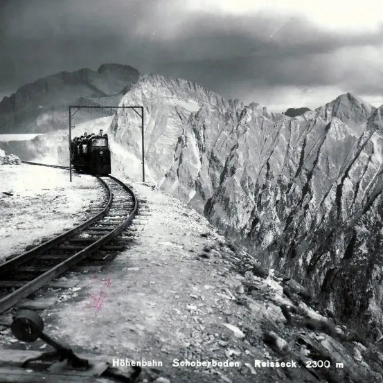 Ein Schwarz-Weiß-Foto zeigt einen Zug, der auf einer Bergbahn mit steilen Hängen und bewölktem Himmel fährt.