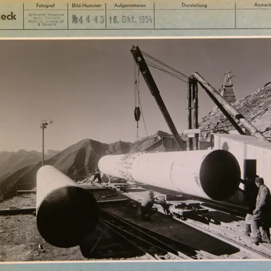 Ein Schwarz-Weiß-Foto vom 16. Oktober 1954, das große Rohre zeigt, die mit Kränen gehandhabt werden. Arbeiter sind auf der rechten Seite sichtbar.