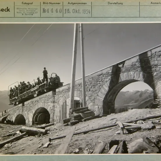 Ein Schwarz-Weiß-Foto einer Brücke mit Menschen, die darauf fahren. Das Bild trägt den Titel 'Wilhelm Wagner' und zeigt die Brücke mit einem Berg im Hintergrund. Das Foto wurde am 16. Oktober 1954 aufgenommen.