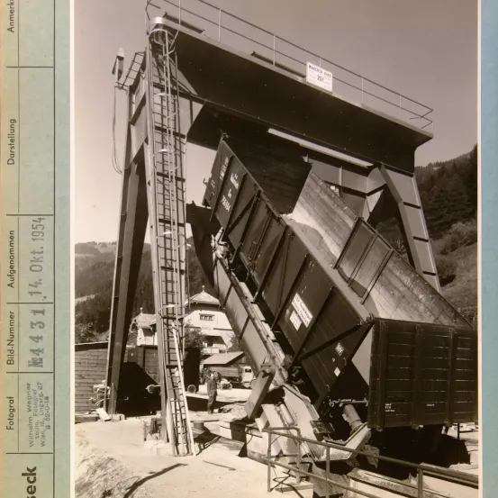 Eine große Industriemaschine ist auf einer Schiene umgekippt, mit einer Person in der Nähe. Ein Schild zeigt 'MACATA'. Im Hintergrund sind Häuser und Bäume zu sehen.