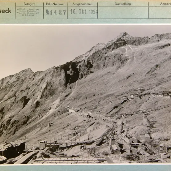 Ein Schwarz-Weiß-Foto eines bergigen Geländes, aufgenommen am 16. Oktober 1954 von Wilhelm Wagner. Das Foto zeigt verschiedene Strukturen, möglicherweise Gebäude und Infrastruktur, eingebettet in die raue Landschaft.