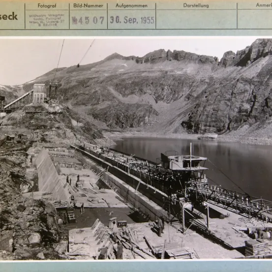 Schwarz-Weiß-Foto einer Staumauer im Bau. Das Bild zeigt Details wie ein Boot, Arbeiter und ein Baufahrzeug. Datum: 30. September 1955.