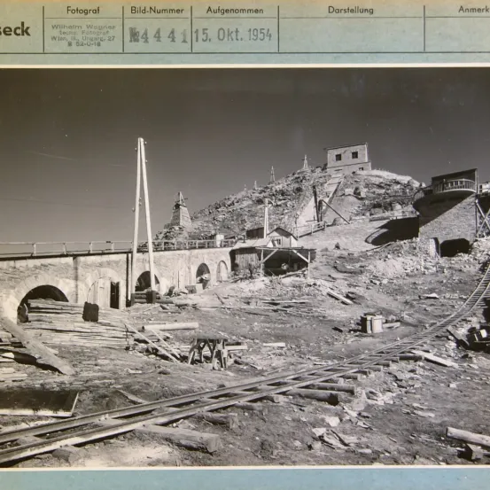 Schwarz-Weiß-Foto einer zerstörten Brücke und verstreutem Schutt. Im Hintergrund ist ein kleines Gebäude und ein Turm zu sehen. Datum: 15. Okt. 1954.