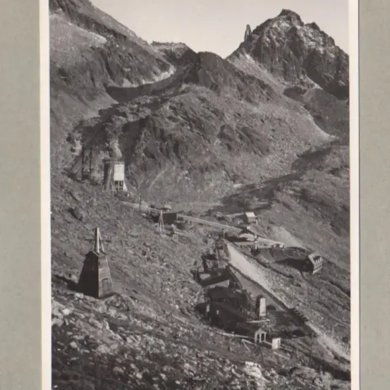 Ein Schwarz-Weiß-Foto einer Gebirgslandschaft mit einer kleinen Struktur am Boden und einem Bergpfad, der zu einem entfernten Gipfel führt.