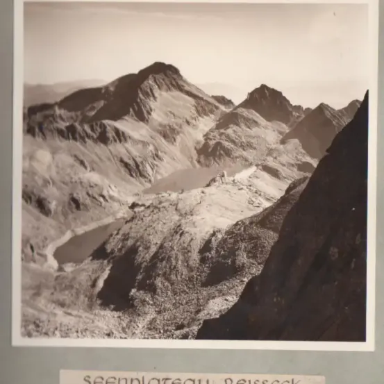 Ein schwarz-weißes Bild zeigt eine Gebirgslandschaft mit rauen Gipfeln, schneebedeckten Bereichen und einem zwischen ihnen gelegenen See. Der Himmel darüber ist klar.