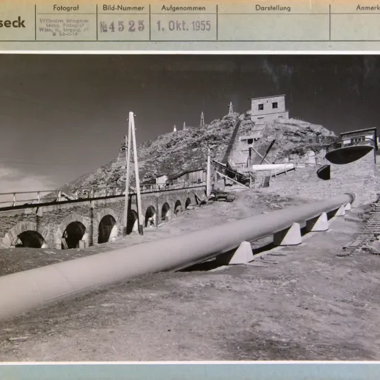 Schwarz-Weiß-Foto mit Bildunterschrift, das eine Baustelle mit einer großen Rohrleitung und einem Gebäude auf einem Hügel zeigt. Das Foto wurde am 1. Oktober 1955 aufgenommen.