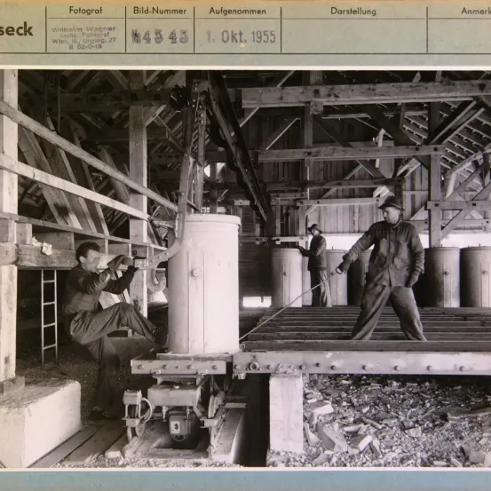 Ein Schwarz-Weiß-Foto von 1955 zeigt zwei Männer, die auf einer Baustelle arbeiten, einer sitzt und der andere steht mit einem Werkzeug. Holzbalken und Tanks sind sichtbar.
