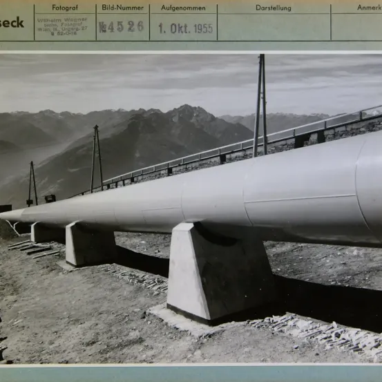 Ein Schwarz-Weiß-Foto zeigt eine Pipeline auf einer Baustelle mit Bergen im Hintergrund. Das Foto ist vom 1. Oktober 1955 und wurde von Wilhelm Wagner aufgenommen.