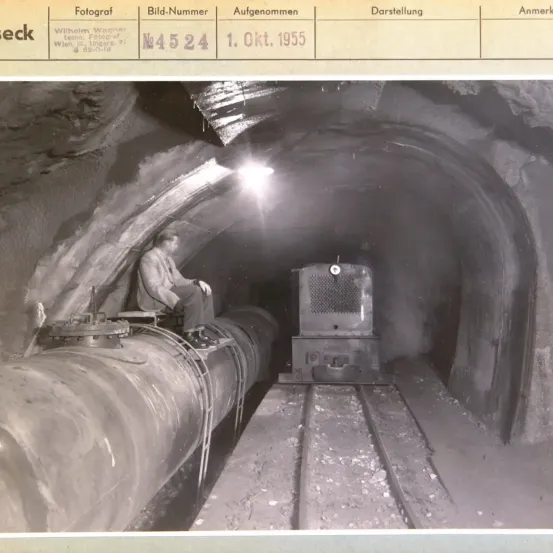 Ein Schwarz-Weiß-Foto eines Mannes, der auf einer Rohrleitung in einem Tunnel sitzt, mit einem Zug auf den Gleisen. Das Foto ist vom 1. Oktober 1955.