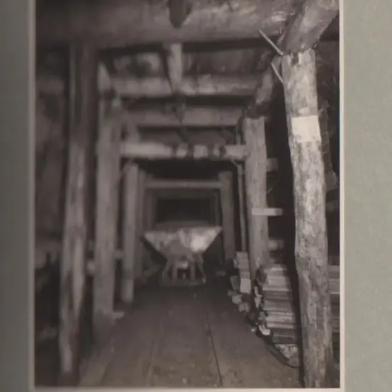 Ein Schwarz-Weiß-Foto zeigt das Innere eines Bergwerkstunnels mit hölzernen Stützbalken und einer räderartigen Struktur in der Mitte.